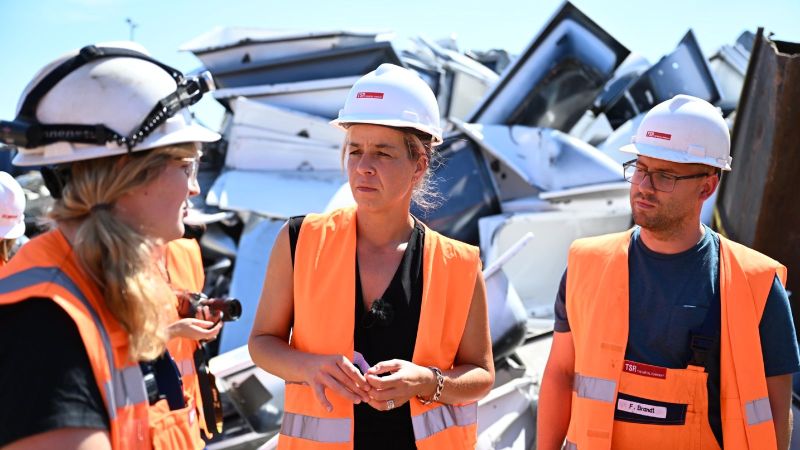 Ministerin Neubaur und zwei weitere Personen mit Helmen auf einem Schrottplatz.