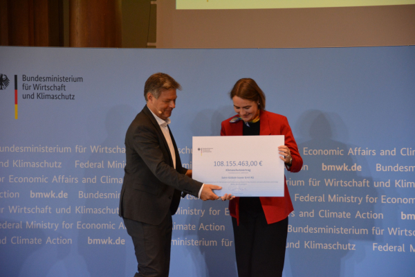 Die Vertreterin der Firma Saint-Gobain ISOVER G+H AG nimmt ein großes Plakat als Symbol für den Klimaschutzvertrag von Robert Habeck entgegen.