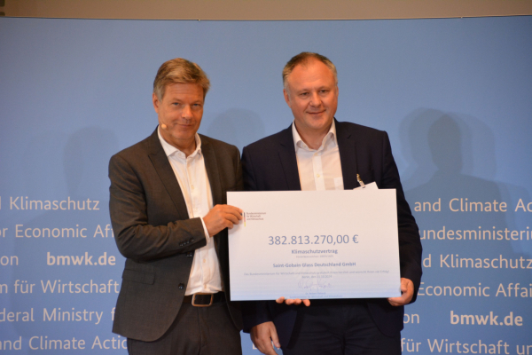 Der Vertreter der Firma Saint-Gobain Glass Deutschland GmbH und Robert Habeck ste-hen nebeneinander und halten ein großes Plakat vor sich, das den Klimaschutzvertrag symbolisiert. 